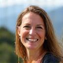 Porträt von Anna aus München, lächelnd vor einer verschwommenen Bergkulisse.